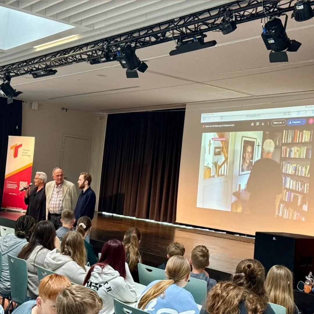 Tils Eltern mit Lehrer vor der Aula, im Hintergrund läuft ein Film über die Geschichte.